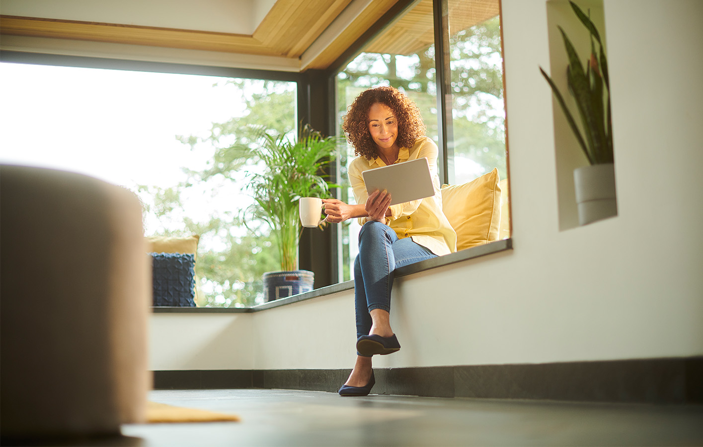Frau auf einer Fensterbank mit großen Fenstern, entspannt mit einer Tasse Kaffee.